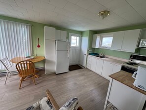 Ett kompakt kök med vita skåp, ett matbord i trä och utsikt över havet genom ett fönster.