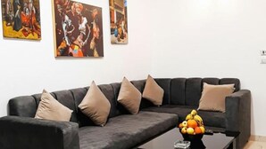 Ein modernes Wohnzimmer mit einem dunkelgrauen Sofa, beigen Kissen und einem Couchtisch mit Früchten. An der Wand hängen drei Gemälde.