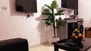 Ein Wohnzimmer mit einem an der Wand befestigten Flachbildfernseher, einem Blumentopf, einem Tisch mit Obstschale und einer Waschmaschine im Hintergrund.