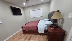 Quarto com cama, mesa de cabeceira, abajur e televisão na parede.