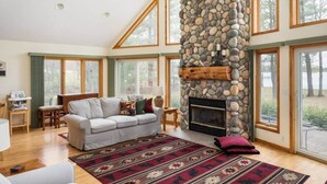A spacious living room with a stone fireplace, large windows, and a cozy seating area. - Lakefront, 5BR Home w/Sandy Beach, Dock & Kayaks (Excelsior Township)