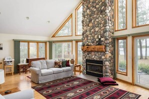 A spacious living room with a stone fireplace, large windows, and a cozy seating area. - Lakefront, 5BR Home w/Sandy Beach, Dock & Kayaks (Excelsior Township)