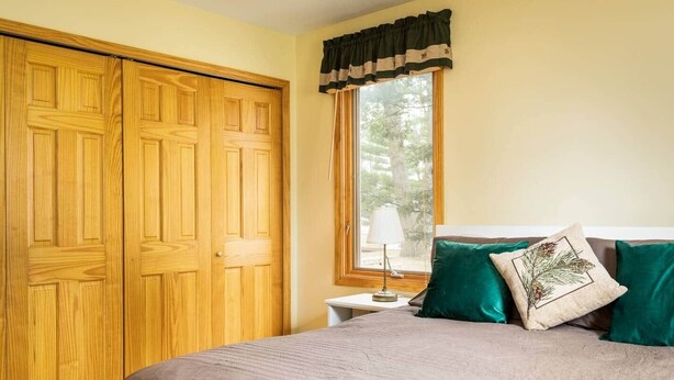 A bedroom with a bed, a nightstand, a window with curtains, and a wooden wardrobe. - Lakefront, 5BR Home w/Sandy Beach, Dock & Kayaks (Excelsior Township)