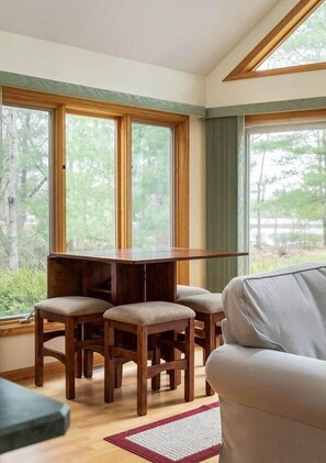 Ein Zimmer mit Holztisch, drei Stühlen, großen Fenstern mit Blick ins Grüne und einem Sofa.