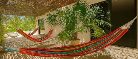 A hammock under a thatched roof, a potted palm tree, and a modern building with a glass door.