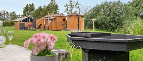 A wooden cabin with a fenced yard, a barbecue grill, and a table with a bottle and flowers.