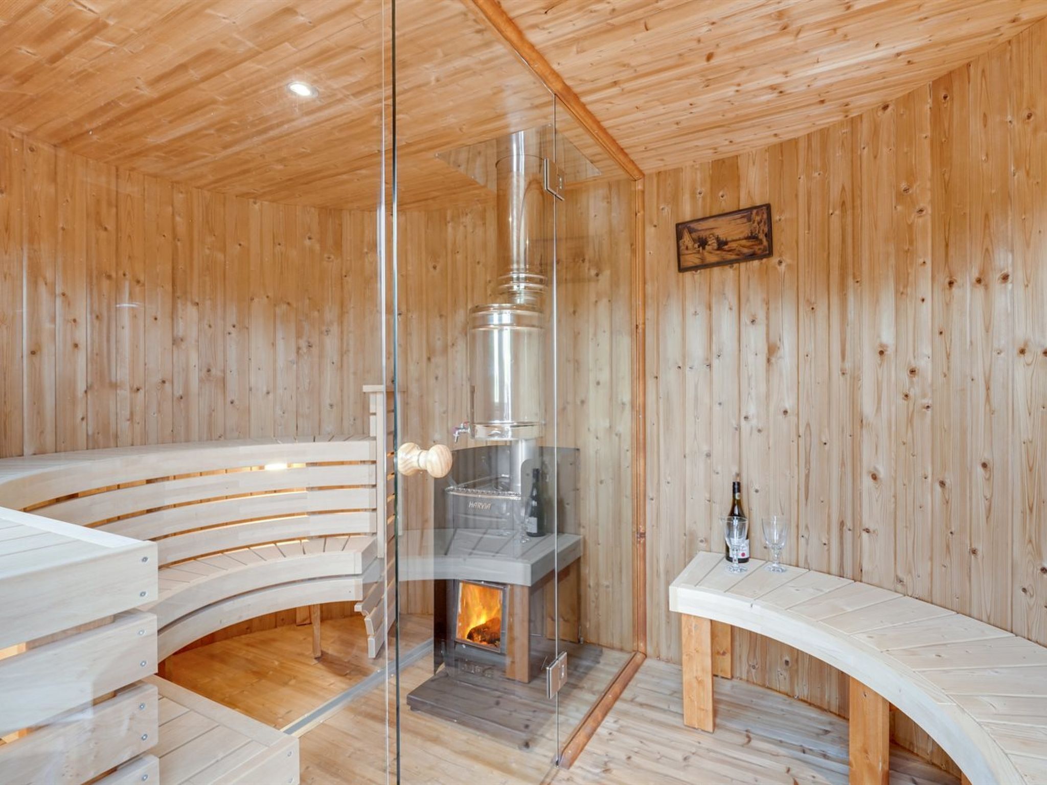 A wooden cabin interior with a glass-enclosed wood stove, seating areas, and a small table with a bottle and glass.