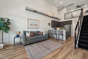 A modern living room with a grey sofa, a coffee table, and a plant. There is a kitchen area with dark cabinets and stainless steel appliances. A staircase with a black railing leads to an upper level. - Red Raider Loft (Lubbock)