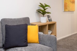 Ein graues Sofa mit dunklem und gelbem Kissen, ein weißer Topfpflanze und ein Holztischchen mit Schalen.