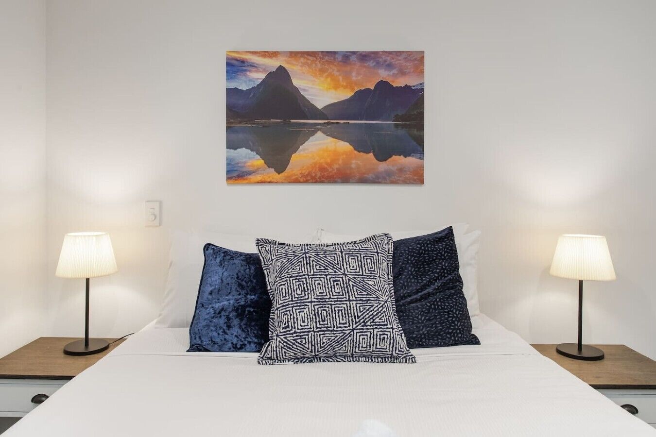 Una cama con ropa de cama blanca, dos mesitas de noche con lámparas, y un cuadro de una montaña y un lago en la pared.