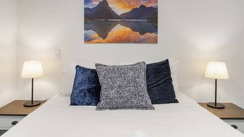 Una cama con ropa de cama blanca, dos mesitas de noche con lámparas, y un cuadro de una montaña y un lago en la pared.