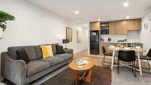 Una sala de estar moderna con un sofá gris, una pequeña mesa redonda y una zona de cocina con armarios de madera y electrodomésticos.