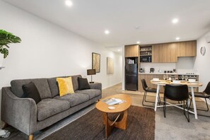 Ein modernes Wohnzimmer mit einem grauen Sofa, einem kleinen runden Tisch und einer Küchenzeile mit Holzschränken und Geräten.