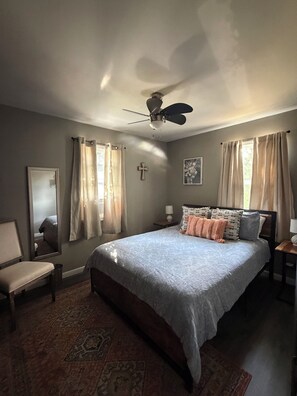 A bedroom with a bed, a chair, a ceiling fan, a cross on the wall, and a window with curtains. - Delightful 3-bedroom house in Rapid City perfect for your getaway (Rapid City)