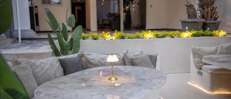 Un espacio exterior con mesas y sillas redondas, rodeado de plantas y vistas al interior.