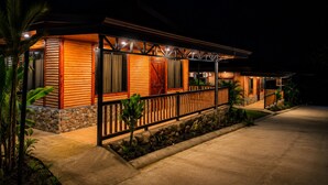A two-story log cabin with a porch, stone foundation, and a well-lit exterior.