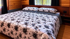 A bedroom with a wooden ceiling, a large bed with floral bedding, an air conditioning unit, and a window with curtains.