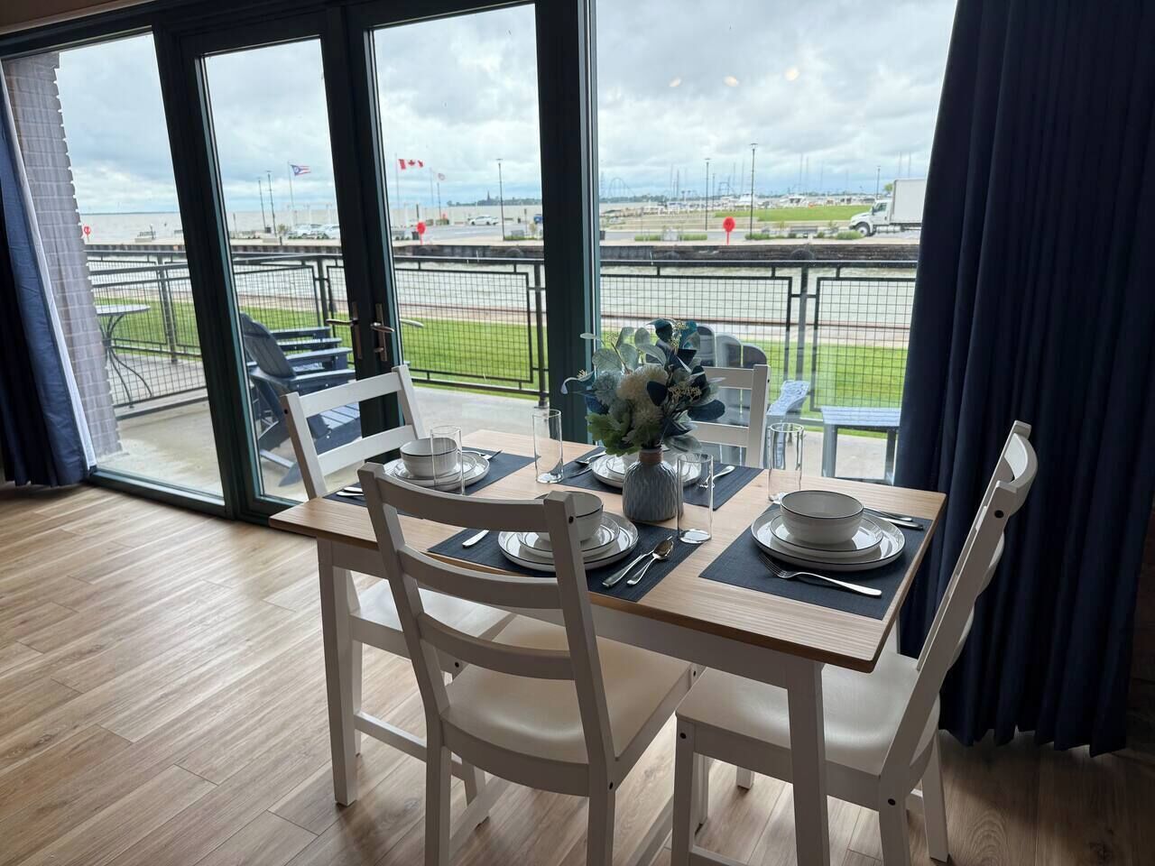 A dining area with a table set for four, a vase with flowers, and a view of a sports field through large windows.