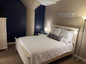 Una cama bien tendida en una habitación con una pared azul oscuro de acento, una mesita de noche con una lámpara, y un cartel sobre la cama.