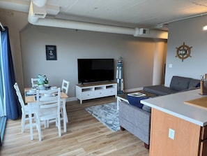 A modern living room with a flat-screen TV, a dining table with chairs, a grey sofa, and a small kitchen area.