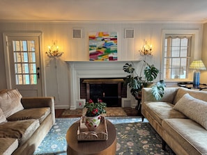 Una sala de estar con chimenea, dos sofás, una mesa de centro con un arreglo floral y un cuadro abstracto grande sobre la repisa.