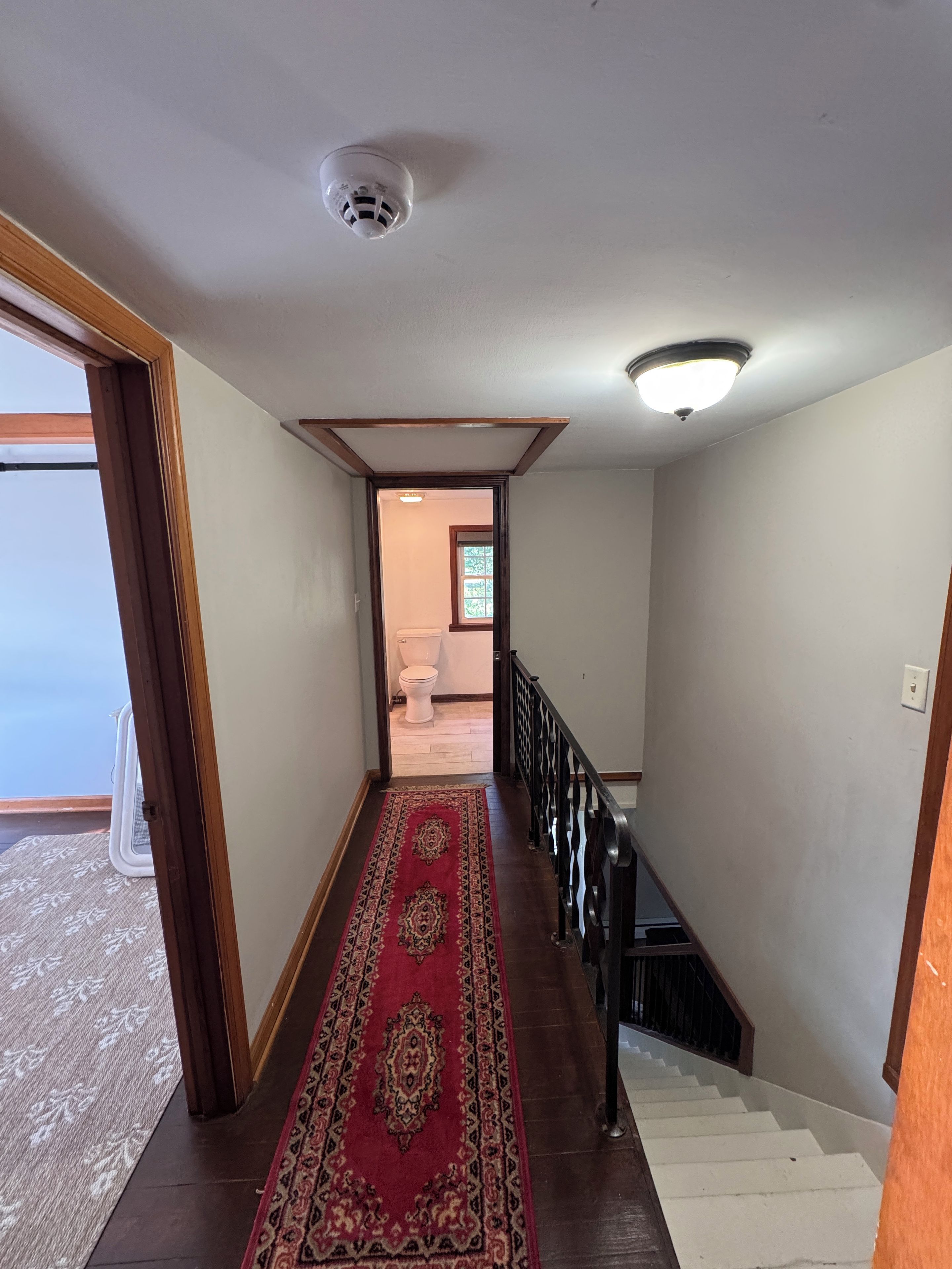Un couloir doté d’un plancher en bois, d’un tapis rouge à motifs, d’un escalier blanc et d’une caméra de surveillance fixée au plafond.