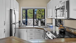 A modern kitchen with white cabinets, a stainless steel refrigerator, a double sink, and a large window offering a view of trees.