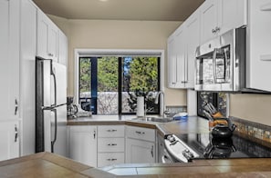 Eine moderne Küche mit weißen Schränken, einem Edelstahl-Kühlschrank, einer doppelten Spüle und einem großen Fenster mit Blick auf Bäume.