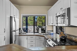 A modern kitchen with white cabinets, a stainless steel refrigerator, a double sink, and a large window offering a view of trees. - River Ridge 420A - Prime Westside River Views | Pool & Hot Tub (Bend)