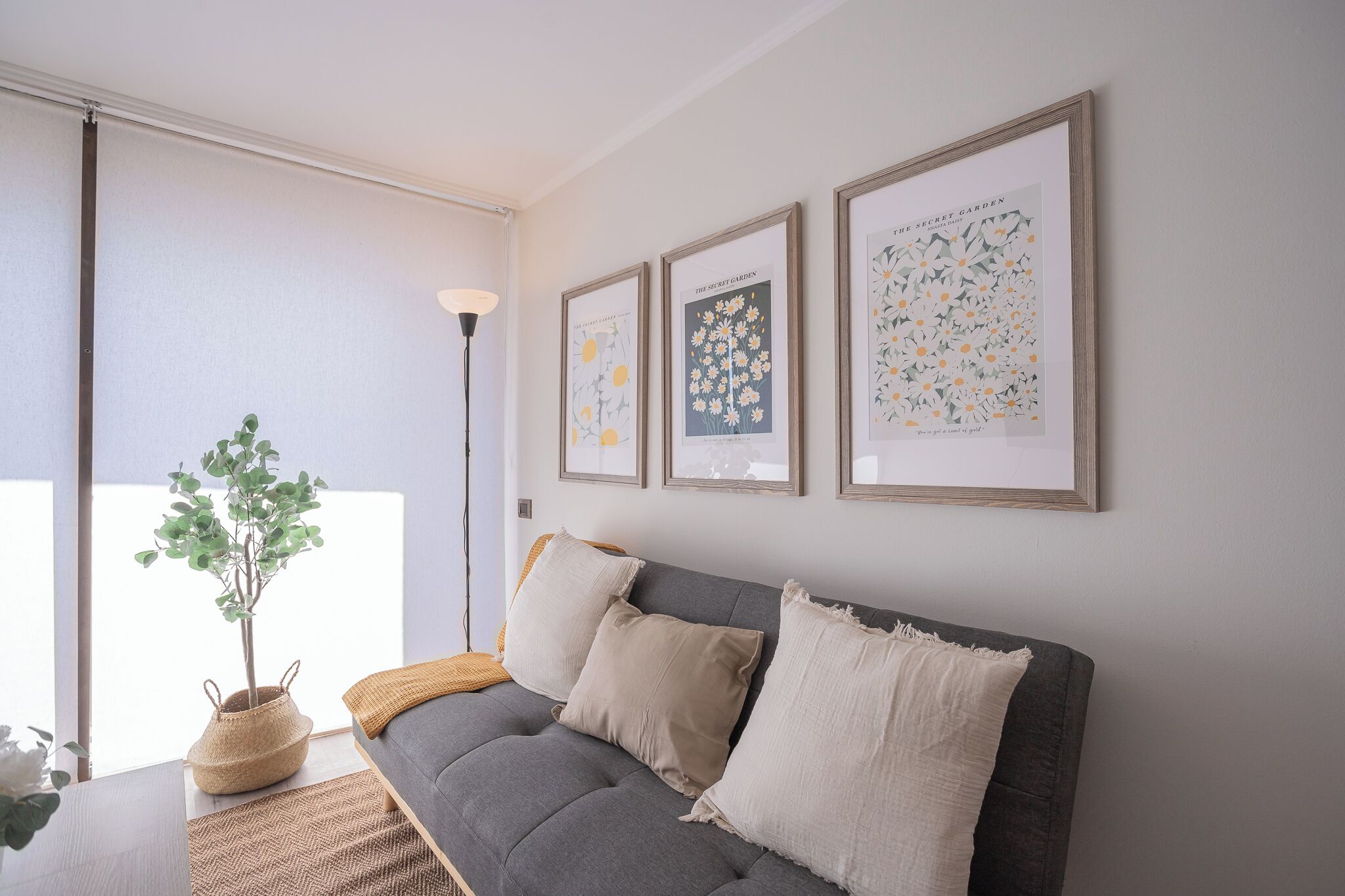 Een moderne woonkamer met een grijze bank, drie ingelijste kunstwerken, een vloerlamp en een potplant.