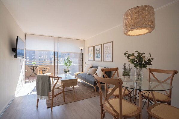 A modern living room with a sofa, coffee table, and chairs, a balcony with a view, and a hanging light fixture. - San Sebastian Apartments By Andes (Las Condes)