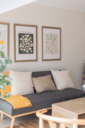 A modern living room with a grey sofa, white and beige cushions, a yellow throw blanket, and a wooden coffee table. There are three framed botanical prints on the wall above the sofa. - San Sebastian Apartments By Andes (Las Condes)