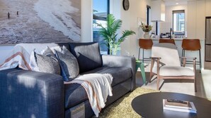 Ein modernes Wohnzimmer mit Schlafsofa, Essbereich mit Stühlen, einer Küche im Hintergrund und einem großen Fenster mit Blick auf den Strand.