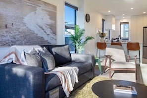 A modern living room with a sofa bed, a dining area with chairs, a kitchen in the background, and a large window with a view of the beach. - Peaceful Merivale Hideaway - Parkside + Parking (Christchurch)