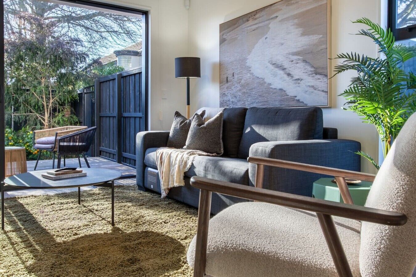 Una sala de estar con un sofá, una mesa de centro y una silla. Hay un ventanal grande, un cuadro en la pared y una planta.