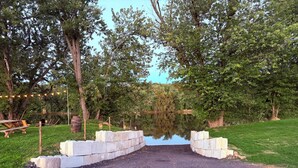 Een grindpad dat leidt naar een waterpartij, omgeven door bomen en een gazon met houten banken en lantaarns.