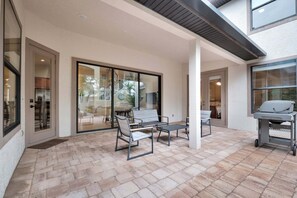 Ein überdachter Patio-Bereich mit Gartenmöbeln, einem Grill und Blick ins Innere durch Glastüren.