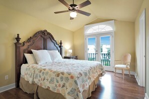Ein Schlafzimmer mit einem großen Bett, einem Deckenventilator, einem Nachttisch, einem Sessel und Blick ins Freie durch eine Tür.