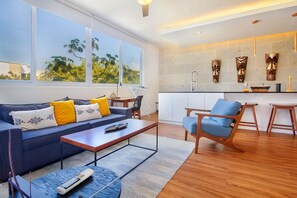 Ein modernes Wohnzimmer mit einer Couch, einem Couchtisch und einem blauen Sessel. Es gibt eine Küchenzeile mit einem Spülbecken und dekorativen Elementen an der Wand.