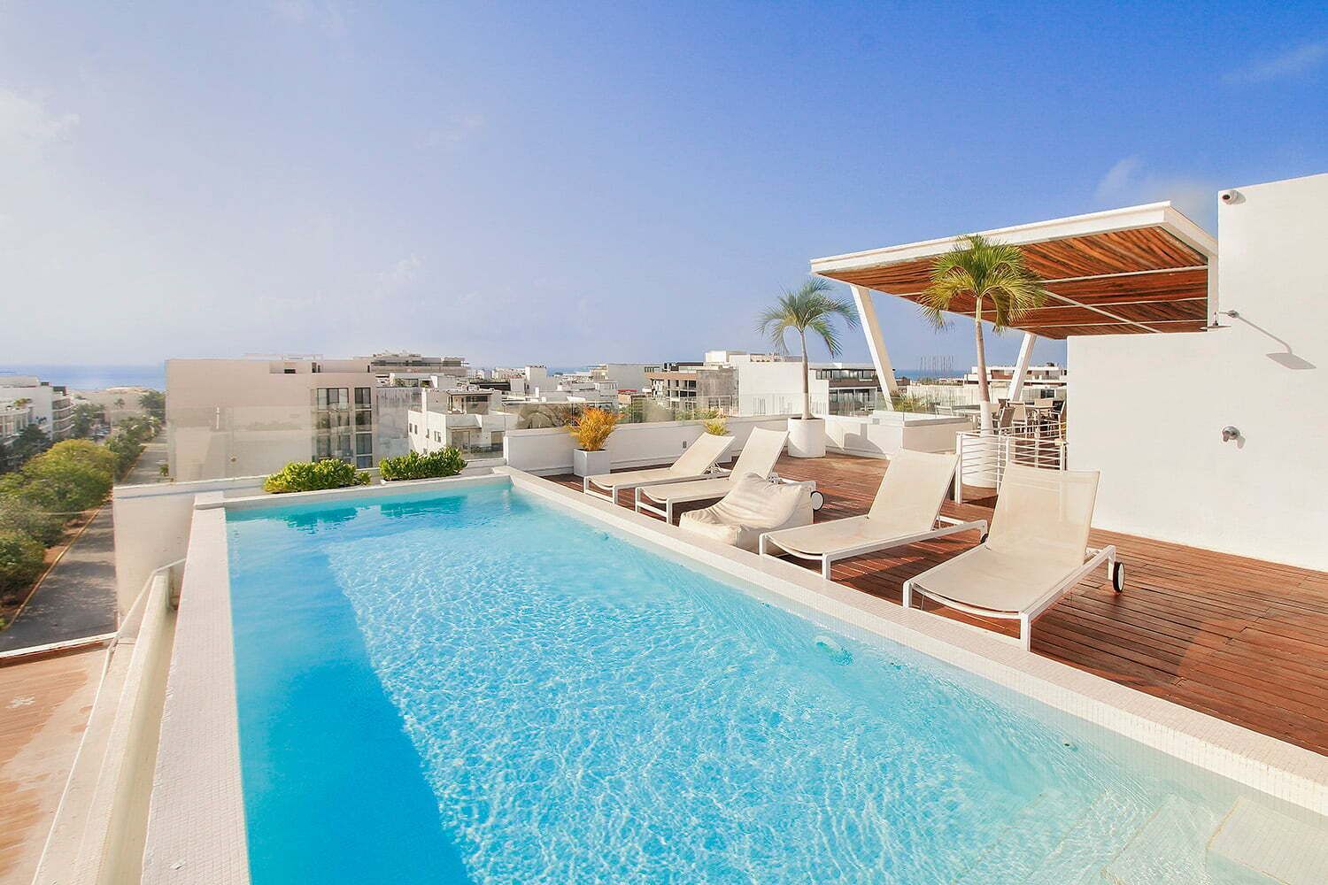 A rooftop infinity pool with lounge chairs and a wooden deck, overlooking a cityscape.