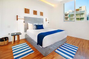 A modern bedroom with a bed, wooden headboard, and a view of buildings through a window. - 2BR Downtown Condo Walk to Coco Beach (Playa Del Carmen)