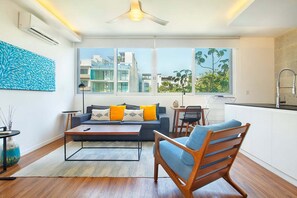 A modern living room with a sofa, coffee table, and wooden chair. There is a kitchen area with a sink and cabinets. Large windows provide a view of the cityscape. - 2BR Downtown Condo Walk to Coco Beach (Playa Del Carmen)