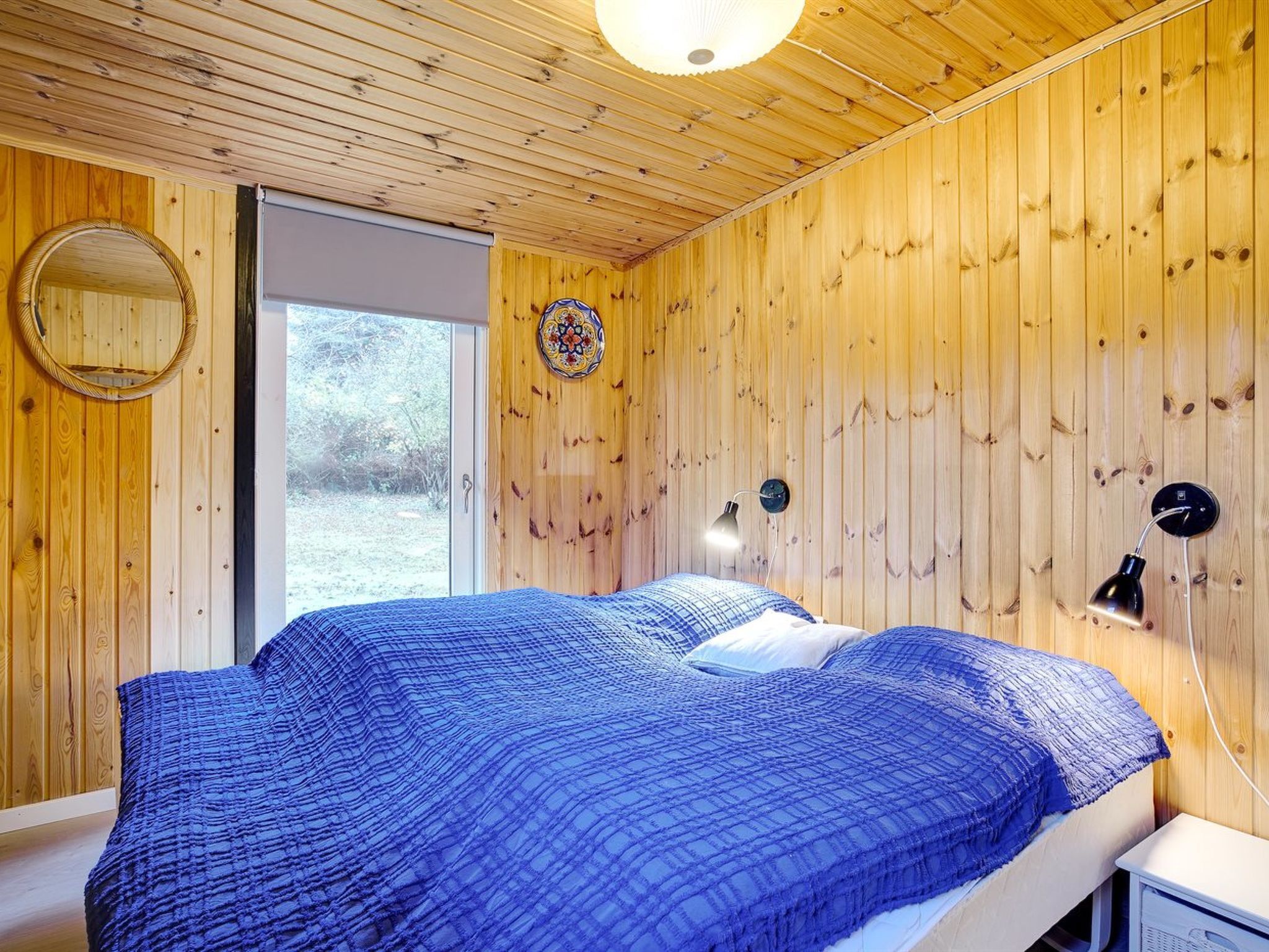 A bedroom with a wooden paneled wall, a bed with blue bedding, a round mirror, a decorative plate, and a window with a view of trees.
