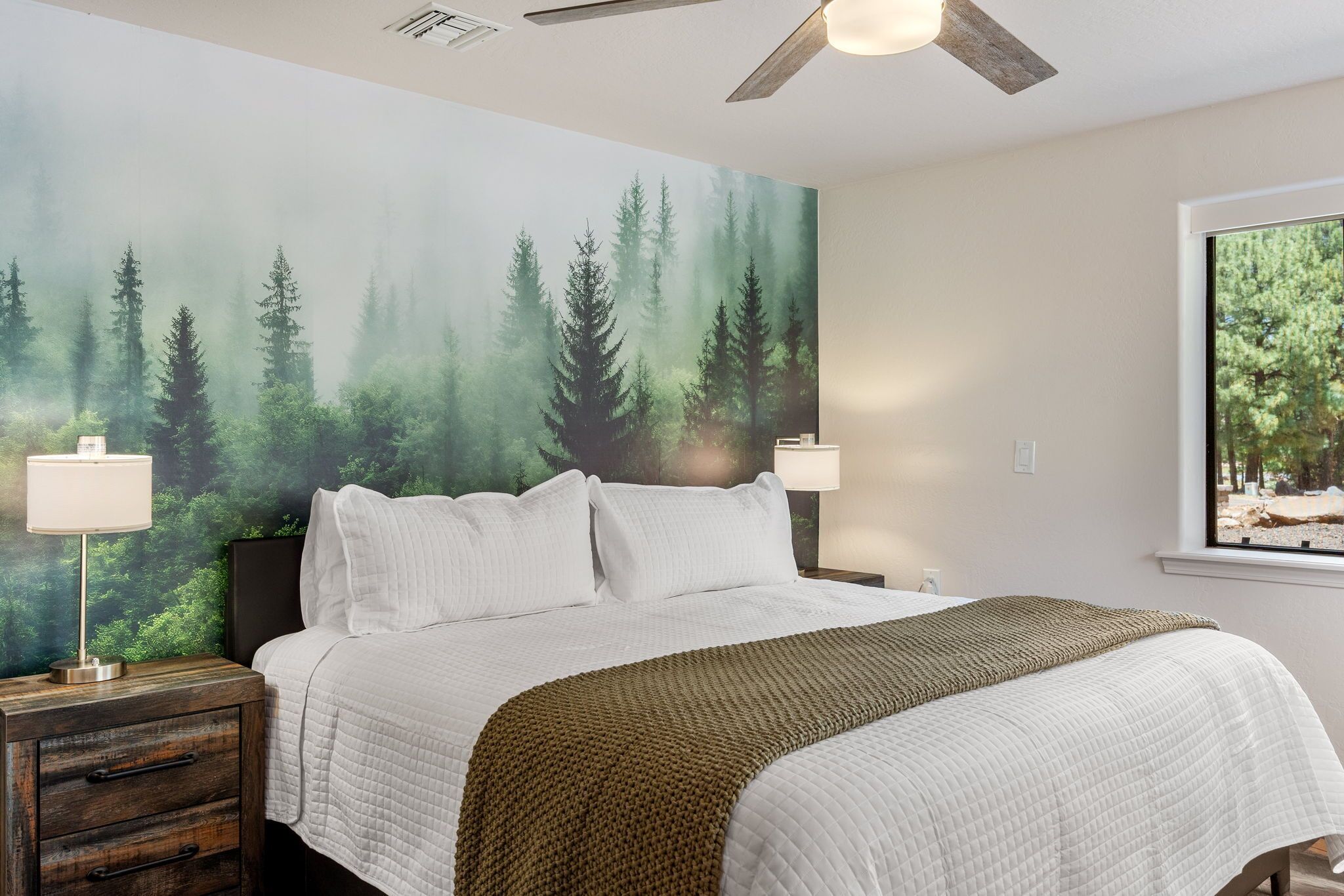 A bedroom with a large bed, two bedside lamps, a wooden nightstand, and a forest-themed wall mural.