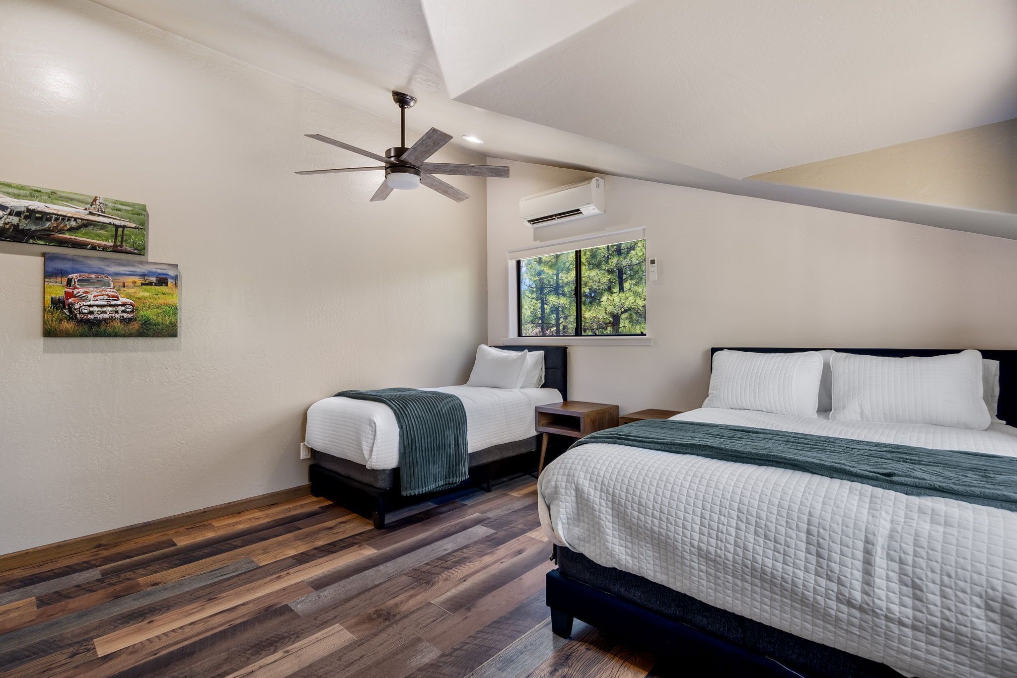 A bedroom with a bed, a smaller bed, a ceiling fan, a window with a view of trees, and artwork on the wall.