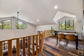 A spacious room with a high ceiling, large windows, a wooden staircase, a dining area with chairs, and a table.