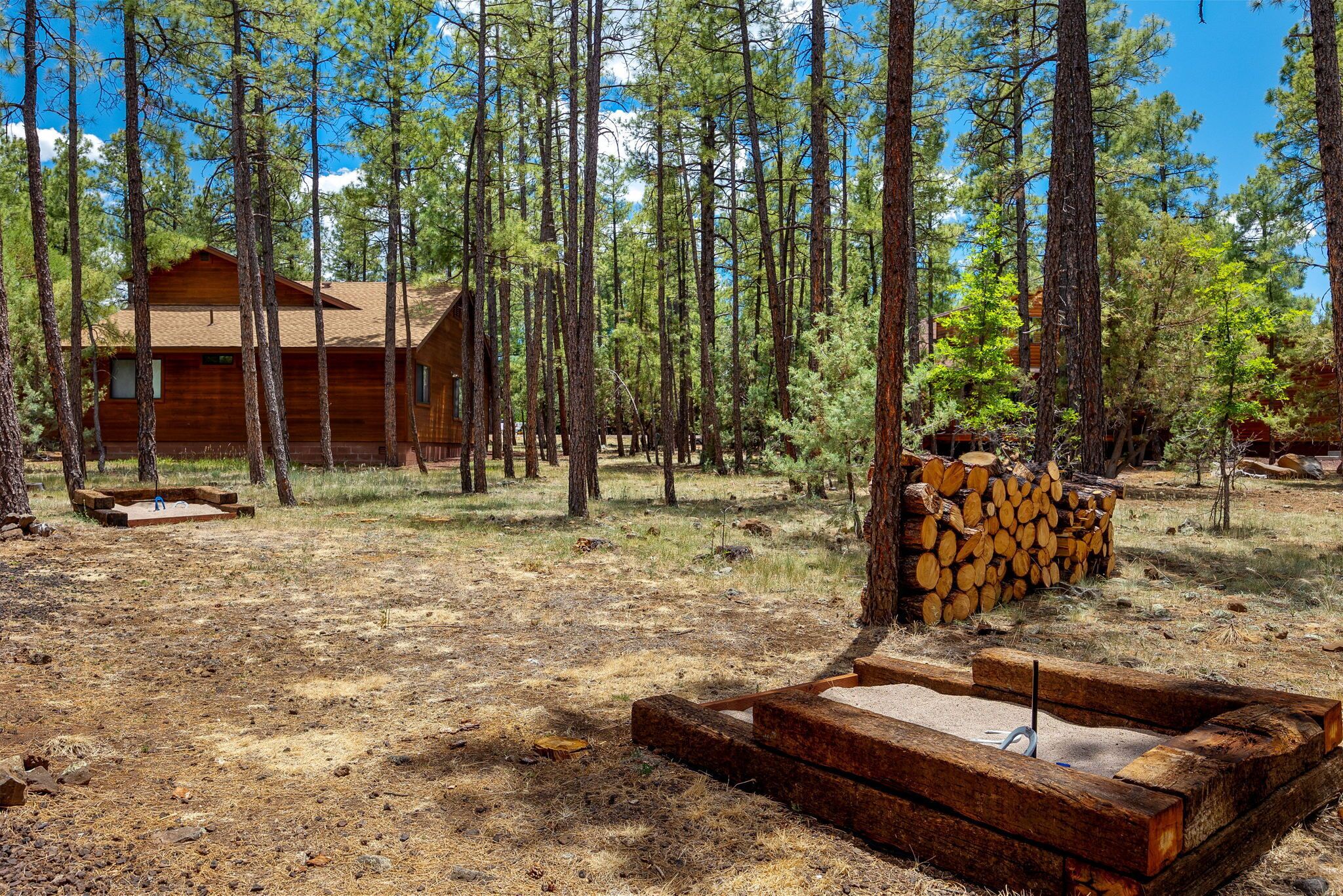 A cabin surrounded by tall pine trees, with a fire pit and stacked firewood nearby.