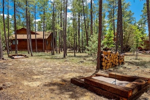 A cabin surrounded by tall pine trees, with a fire pit and stacked firewood nearby. - RainbowLake-HotTub-GameRoom-Kayaks-Horseshoes-EVCharge-Firepit-Pickleball-7Beds (Pinetop-Lakeside)