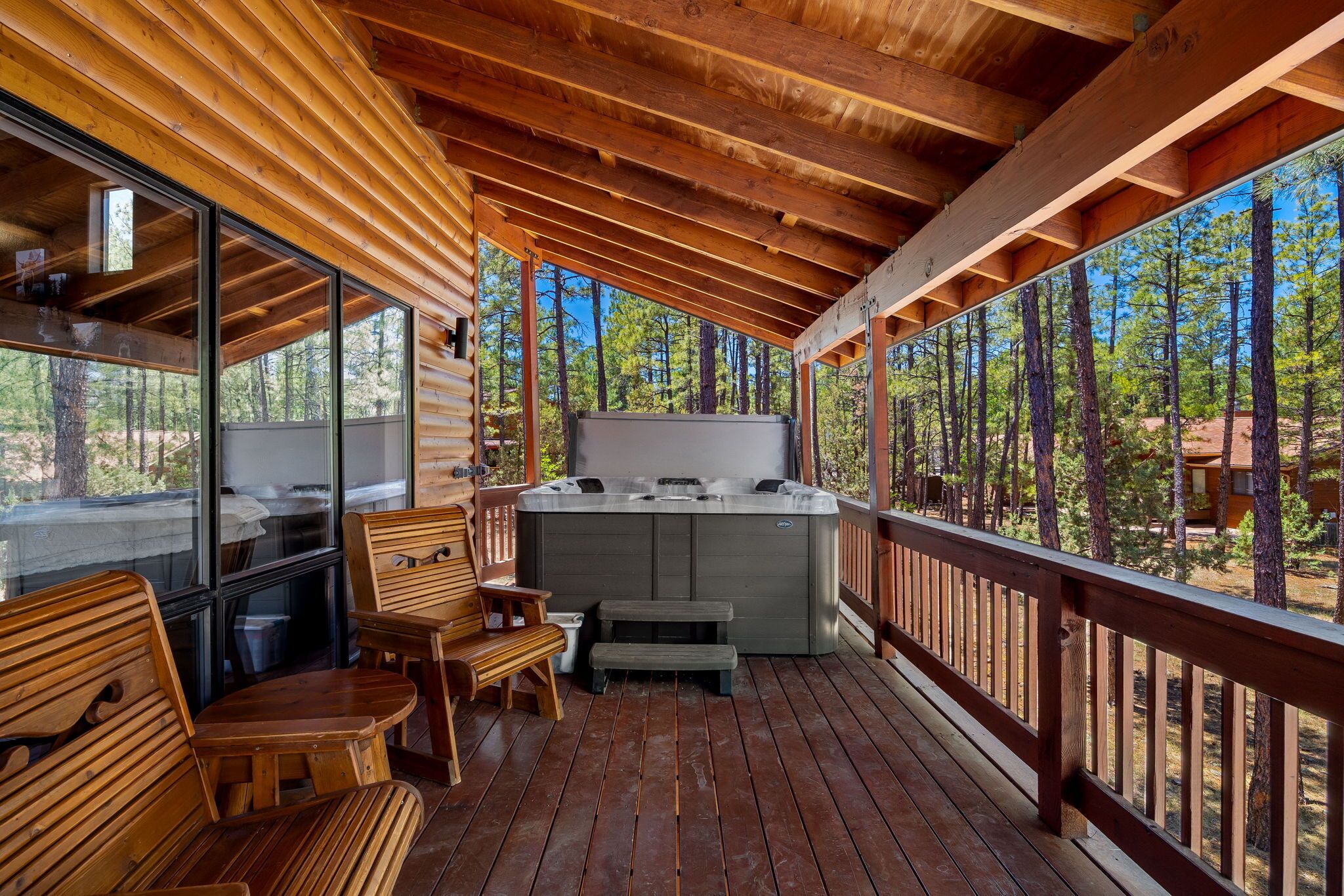 A wooden deck with a hot tub, wooden furniture, and a view of a forest.