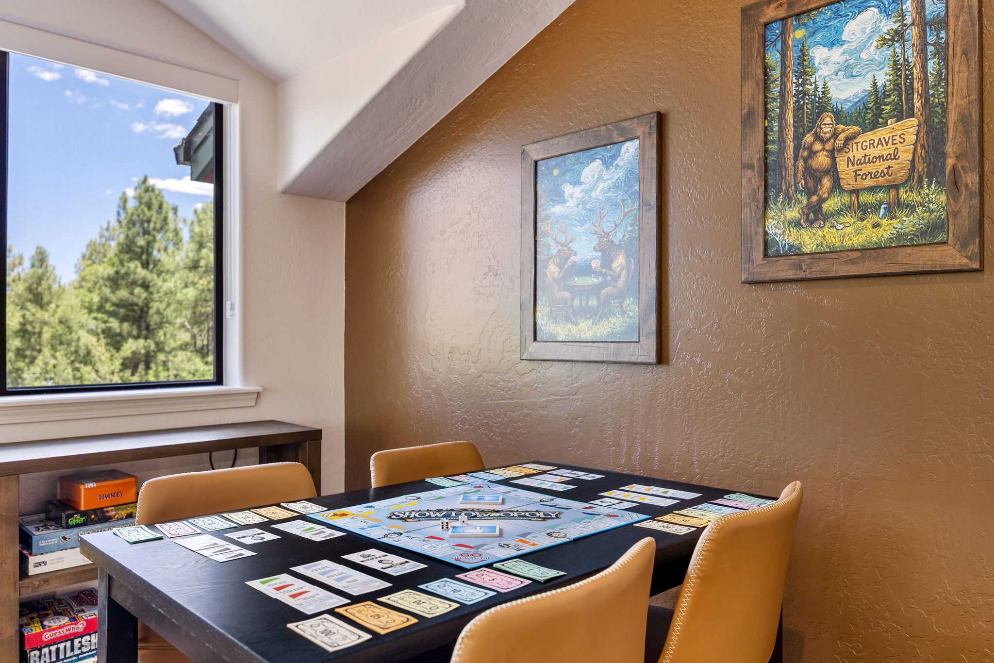 A dining area with a board game set up on a table, framed artwork on the wall, and a window with a view of trees.